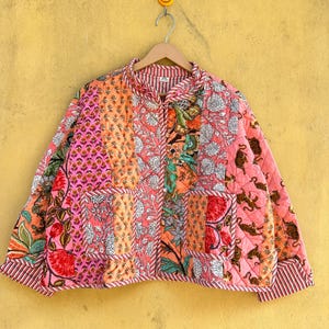 Può includere: Giacca trapuntata con design patchwork nei toni del rosa, arancione e verde. La giacca presenta varie stampe floreali e animali, un collo alto e un bordo a righe. Appesa a una gruccia di legno.