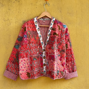 May include: A patchwork quilted jacket in red and pink hues, featuring floral patterns. The jacket has a V-neck, button closure, and striped trim. It is displayed on a wooden hanger, showcasing its design and details. The jacket's measurements are in centimeters.