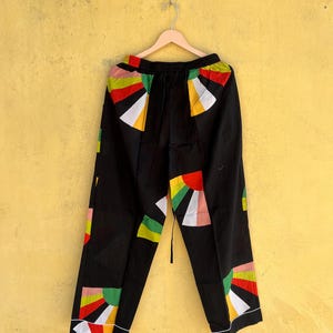 May include: Black pants with a colorful geometric design featuring red, yellow, green, and white shapes. The pants are hanging on a wooden hanger against a yellow wall. The design is on the front and back of the pants.