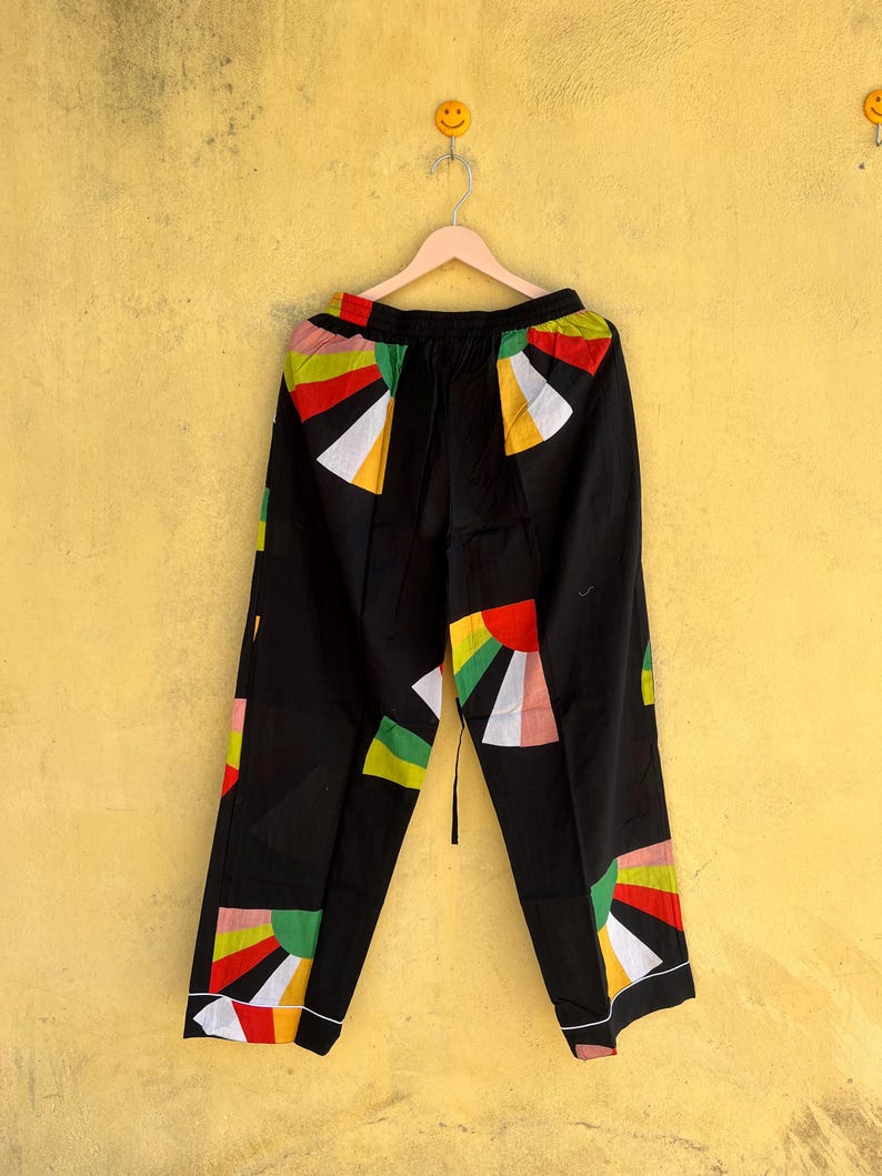 May include: Black pants with a colorful geometric design featuring red, yellow, green, and white shapes. The pants are hanging on a wooden hanger against a yellow wall. The design is on the front and back of the pants.