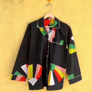 May include: Black pajama top with a colorful geometric design. The shirt has a white trim and button closure. The design includes red, yellow, green, and white shapes. The shirt is hanging on a wooden hanger against a yellow wall.
