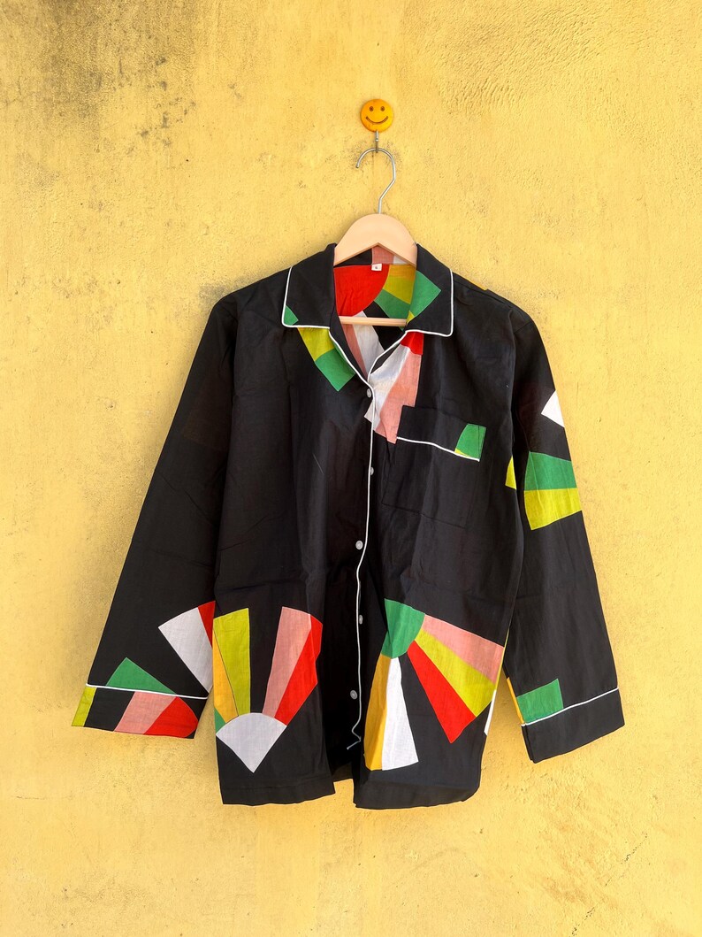 May include: Black pajama top with a colorful geometric design. The shirt has a white trim and button closure. The design includes red, yellow, green, and white shapes. The shirt is hanging on a wooden hanger against a yellow wall.