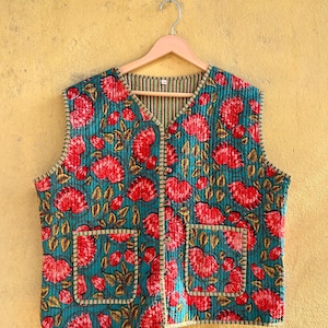 May include: A teal quilted vest with a vibrant floral pattern in red and gold. The vest features two front pockets and a striped lining. It is displayed on a wooden hanger against a yellow background.