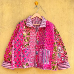 May include: A pink patchwork jacket with floral and geometric designs. The jacket has a collared neckline, button-up front, and two front pockets. The cuffs and collar are trimmed with pink and white striped fabric. The jacket is hanging on a wooden hanger.