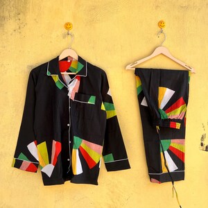 May include: Black pajama set with a colorful geometric design. The long-sleeved top has a button-down front and white trim. The pants have an elastic waistband and a matching pattern. The set is hanging on a wooden hanger against a yellow wall.