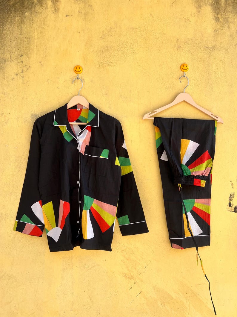 May include: Black pajama set with a colorful geometric design. The long-sleeved top has a button-down front and white trim. The pants have an elastic waistband and a matching pattern. The set is hanging on a wooden hanger against a yellow wall.
