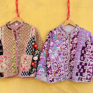 May include: Two quilted jackets with floral and geometric patterns hang against a yellow backdrop. One is tan with red and black accents, the other is purple with white floral designs. Both have pockets and are on wooden hangers.