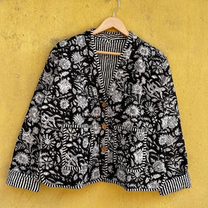 Könnte beinhalten: Schwarz-weiß bedruckte Jacke mit Holzknöpfen und zwei Vordertaschen. Die Jacke hat ein gestreiftes Futter und Manschetten. Sie hängt an einem Holzkleiderbügel vor einer gelben Wand.
