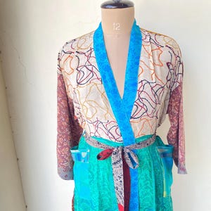 Op de afbeelding: Een kleurrijke kimono met een crèmekleurige top met een patroon van rode, oranje en zwarte lijnen. De kimono heeft een felblauwe kraag en een turquoise rok. Een patroonriem met een rood accent sluit de taille.