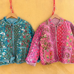 May include: Two quilted jackets with floral patterns hang against a yellow wall. One is teal with white and red flowers, the other is pink with a mix of floral designs. Both have pockets and striped trim.