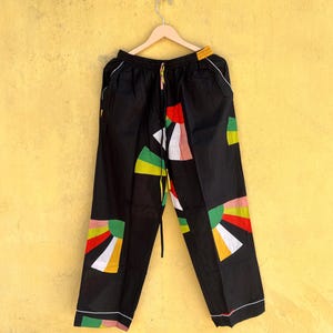 May include: Black pants with colorful geometric designs in red, yellow, green, and white. The pants have a drawstring waist and white trim along the edges. The design is a mix of abstract shapes and sunburst patterns.