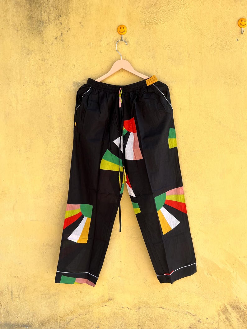 May include: Black pants with colorful geometric designs in red, yellow, green, and white. The pants have a drawstring waist and white trim along the edges. The design is a mix of abstract shapes and sunburst patterns.