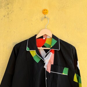 May include: Black long-sleeve shirt with a colorful geometric design on the collar and pocket. The shirt has white trim and buttons. The shirt is hanging on a wooden hanger against a yellow wall.