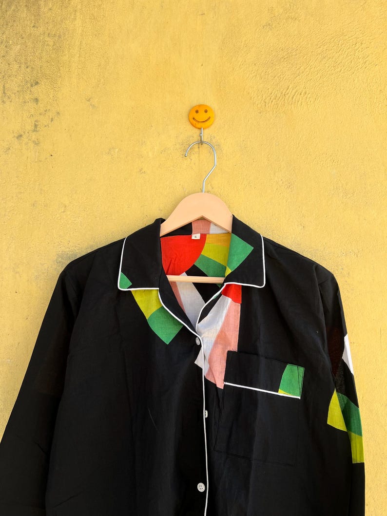 May include: Black long-sleeve shirt with a colorful geometric design on the collar and pocket. The shirt has white trim and buttons. The shirt is hanging on a wooden hanger against a yellow wall.