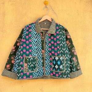 Op de afbeelding: Een gewatteerde jas met een patchwork ontwerp. De jas heeft een mix van bloemen-, zigzag- en streepmotieven in groen, roze en wit. Het heeft twee voorzakken en een gestreepte kraag en manchetten. De jas hangt aan een houten hanger.