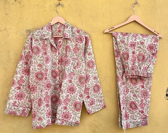 May include: A two-piece pajama set with a floral pattern. The set includes a button-down shirt and matching pants, both in a cream color with a repeating pattern of pink flowers and green leaves. The pajamas are hanging on wooden hangers against a yellow wall.