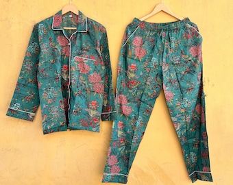 May include: Teal-colored pajama set with a floral pattern in red and blue. The set includes a button-down shirt with white trim and matching pants. The pajamas are hanging on wooden hangers against a yellow wall.