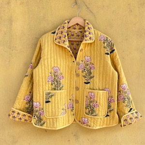 May include: A yellow quilted jacket featuring a pink and green floral design. The jacket has a collared neckline, two front pockets, and button fastenings. The cuffs and collar are lined with a matching floral pattern.