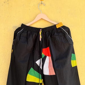 May include: Black pants with a colorful geometric pattern. The design includes red, green, yellow, and white shapes. The pants have a drawstring waist and white trim along the sides.