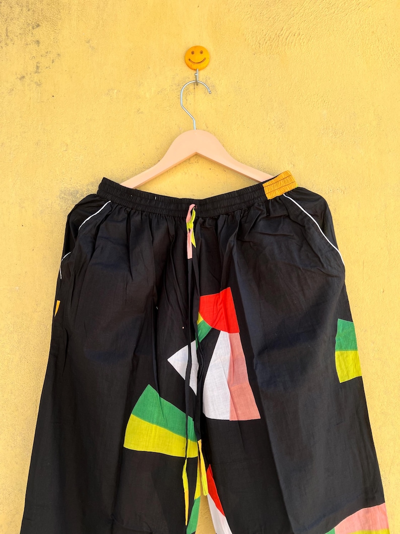 May include: Black pants with a colorful geometric pattern. The design includes red, green, yellow, and white shapes. The pants have a drawstring waist and white trim along the sides.