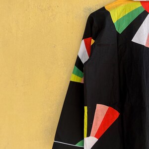 May include: Black button-up shirt with a colorful geometric design. The shirt features a white collar and cuffs, and the sleeves are adorned with red, yellow, green, and white shapes. The shirt is hanging against a yellow background.
