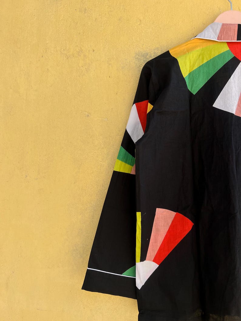 May include: Black button-up shirt with a colorful geometric design. The shirt features a white collar and cuffs, and the sleeves are adorned with red, yellow, green, and white shapes. The shirt is hanging against a yellow background.