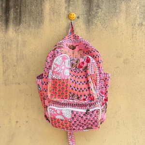 Könnte beinhalten: Ein gesteppter Stoffrucksack mit einem leuchtend rosa Patchwork-Design und verschiedenen Blumen- und Pflanzenmustern. Die Tasche verfügt über ein Hauptfach, eine Reißverschlusstasche auf der Vorderseite und einen Tragegriff, alles mit sichtbaren Nähten.