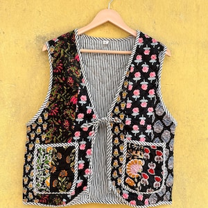 May include: A quilted sleeveless waistcoat featuring a black base with multi-coloured floral block prints, two front patch pockets, and a black and white striped trim along the edges and interior lining, secured with a thin fabric tie at the centre.