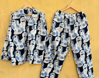 Puede incluir: Un conjunto de pijama de dos piezas con un patrón repetido de perros blancos con detalles azules sobre un fondo azul marino. El conjunto incluye una camisa con botones y pantalones a juego, ambos colgados en perchas de madera.