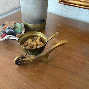 May include: A small, antique brass wheelbarrow-shaped incense burner. The bowl is filled with amber-colored resin. The wheelbarrow has a green and brass finish. The burner is on a wooden surface.