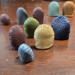 May include: A selection of leather thimbles in shades of brown, green, blue, and tan. Each thimble features a distinct texture and stitching. The thimbles are displayed on a wooden surface.