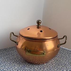 May include: A copper pot with a lid, featuring brass handles and a small brass knob on top. The pot has a rounded shape and a decorative pattern on the lower half. A small rectangular brass plate is affixed to the front.