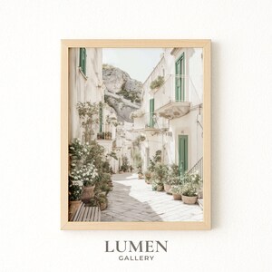 May include: Framed print of a sunlit Italian street scene. White buildings with green shutters and balconies line the cobblestone path. Potted plants and flowers add color. The print is in a light wood frame. The text "LUMEN GALLERY" is at the bottom.
