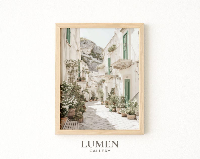 May include: Framed print of a sunlit Italian street scene. White buildings with green shutters and balconies line the cobblestone path. Potted plants and flowers add color. The print is in a light wood frame. The text "LUMEN GALLERY" is at the bottom.