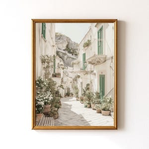 May include: A framed print of a sunlit European street scene. White buildings with green doors and shutters line a cobblestone street. Potted plants and flowers add color. A mountain is visible in the background. The frame is gold.