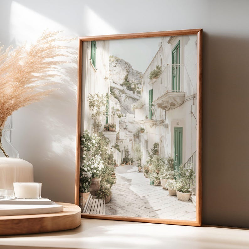 May include: A framed print of a sunlit Italian street scene. The image features white buildings with green doors and balconies, a stone-paved street, and potted plants. The frame is made of wood, and the overall aesthetic is bright and airy.