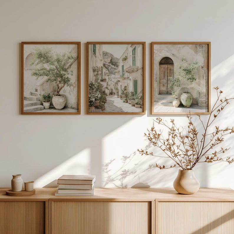 May include: Three framed art prints depicting Italian village scenes. Each print is in a light brown wooden frame. The prints feature neutral tones, with images of buildings, plants, and doorways. The artwork is displayed above a wooden cabinet.