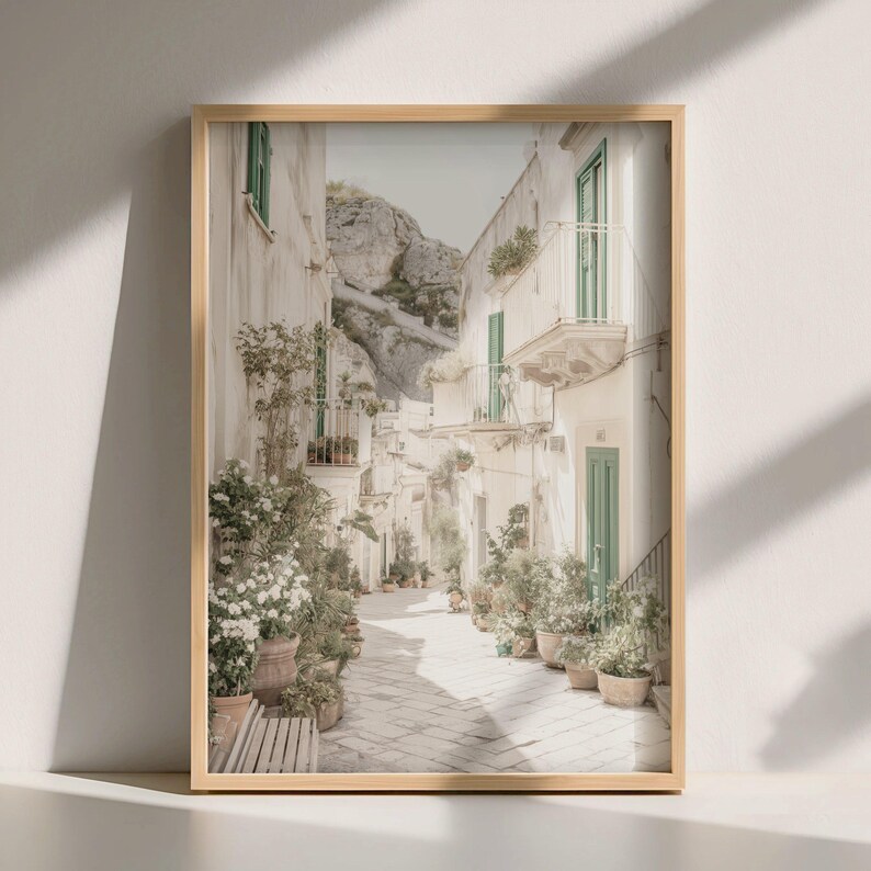 May include: A framed print of a narrow European street scene. White buildings with green shutters and balconies line the cobblestone path. Potted plants and a bench add to the charm. A large rock formation is visible in the background.