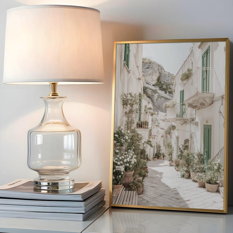 May include: A clear glass table lamp with a white lampshade and gold accents sits next to a framed print. The print depicts a narrow street lined with white buildings, green shutters, and potted plants. The lamp rests on a stack of magazines.