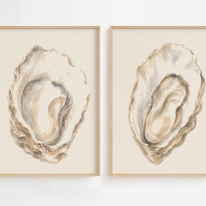 May include: Two framed wall art prints featuring watercolour illustrations of oyster shells. Each print displays a single, detailed oyster shell in shades of beige, tan, and soft grey against a light cream background, enclosed in a light wood frame.