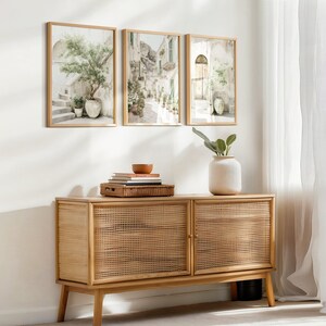 May include: Three framed landscape prints depicting Italian scenes hang above a light wood cabinet. The cabinet has woven cane doors and a minimalist design. A white vase with greenery and a small bowl sit on top of the cabinet.
