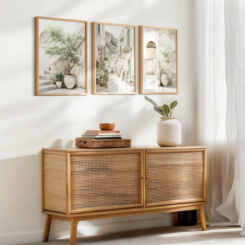 May include: Three framed landscape prints depicting Italian scenes hang above a light wood cabinet. The cabinet has woven cane doors and a minimalist design. A white vase with greenery and a small bowl sit on top of the cabinet.