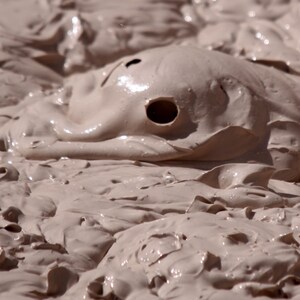 May include: Close-up of a light brown, bubbling mud pot. The mud has a thick, creamy consistency with a few small holes and a smooth, reflective surface. The image captures the texture and movement of the mud.
