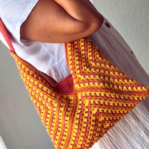 May include: A handmade crochet shoulder bag in a vibrant yellow and orange color scheme. The bag features a granny square design and a solid orange strap. The bag is held by a person wearing a white dress.