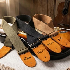 Puede incluir: Tres correas de guitarra en verde oliva, negro y beige, cada una con una pieza final de cuero personalizada. Las correas descansan sobre una guitarra eléctrica con acabado sunburst, con una guitarra acústica al fondo.