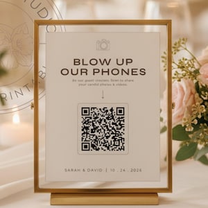 Könnte beinhalten: Ein goldfarbener Rahmen mit dem Text "BLOW UP OUR PHONES" und einem QR-Code. Das Schild steht auf einer weißen Tischdecke, mit einem Blumenarrangement und Kerzen im Hintergrund. Das Schild enthält auch die Namen "SARAH & DAVID" und das Datum "10.24.2026".