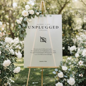 Può includere: Un cartello per la cerimonia nuziale su un cavalletto dorato, circondato da rose bianche e verde. Il cartello recita "Benvenuti alla nostra cerimonia unplugged" con un simbolo di divieto di fotocamere. Il cartello incoraggia gli ospiti a essere presenti.
