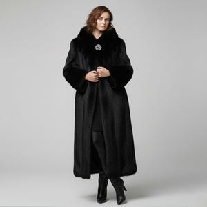 Könnte beinhalten: Ein langer, schwarzer Pelzmantel mit Kapuze und dekorativer Brosche am Ausschnitt. Der Mantel hat lange Ärmel mit Pelzbündchen und eine taillierte Passform. Das Model trägt schwarze Stiefel. Ein luxuriöses Oberbekleidungsstück.