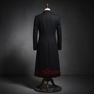 May include: A black, long coat displayed on a mannequin. The coat features red floral embroidery along the hem and cuffs. The coat is a formal style, with a tailored fit and a classic design.