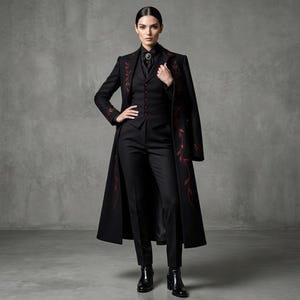 Puede incluir: Un conjunto de traje negro con un abrigo largo con bordado floral rojo, un chaleco y pantalones a medida. El atuendo se completa con una camisa negra y botas. Un broche adorna la camisa.